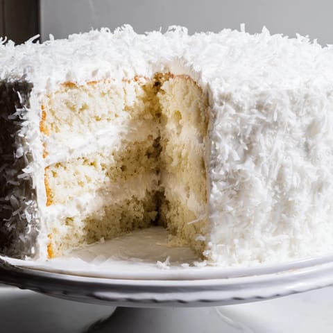 Tall slices of homemade coconut cake reveal fluffy layers and snowy shredded coconut frosting on a marble platter.