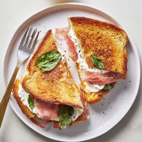 Two halves of a warm Crispy Prosciutto & Burrata Grilled Cheese rest beside fresh basil on a rustic wooden board.