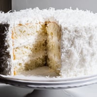 Tall slices of homemade coconut cake reveal fluffy layers and snowy shredded coconut frosting on a marble platter.